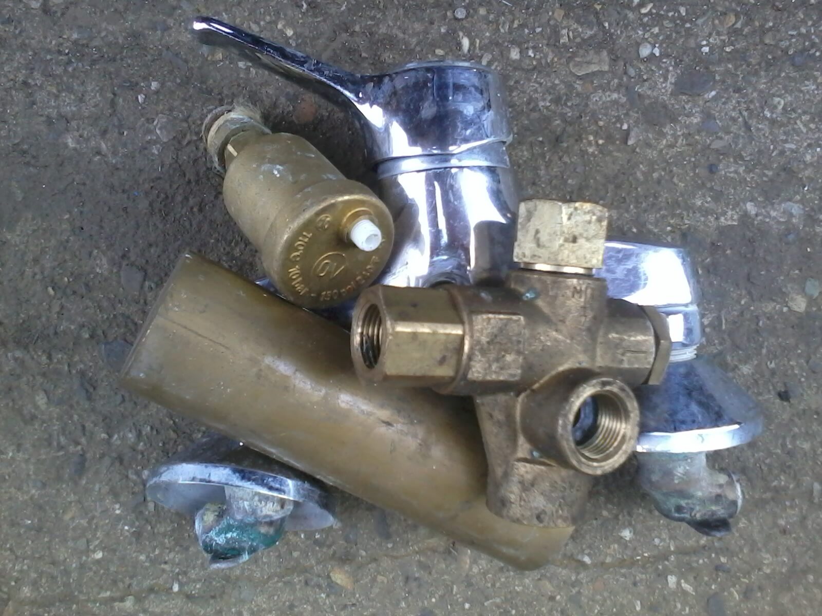 Verschiedene ausgediente Sanitärarmaturen und Metallteile aus Messing und Chrom liegen gesammelt auf einem grauen Betonboden.