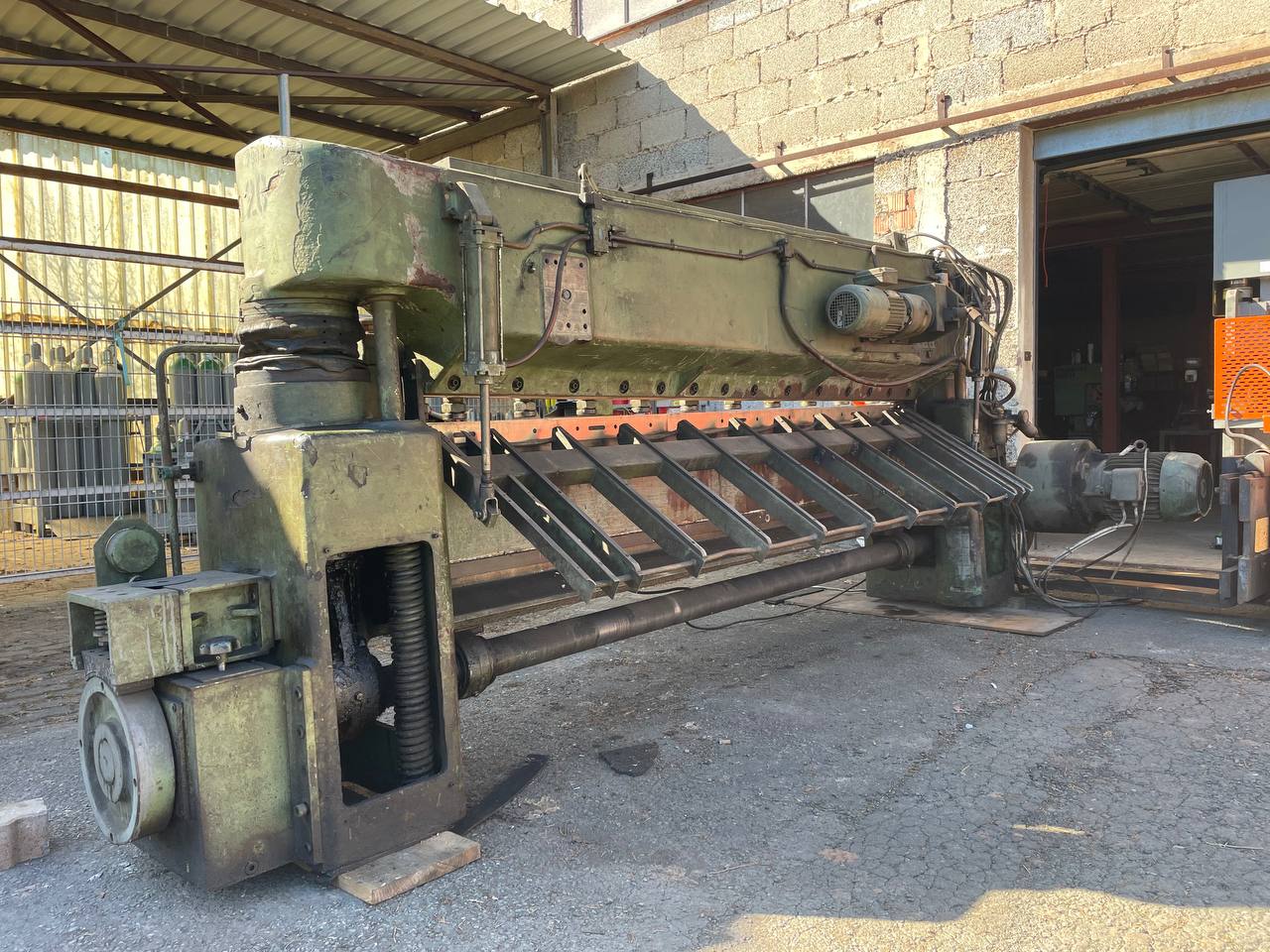 Eine große, massiv gebaute industrielle Metallschere in olivgrüner Farbe steht auf einem Asphaltboden im Freien vor einem Werkstattgebäude. Die Maschine weist Gebrauchsspuren und sichtbare mechanische Komponenten wie Motoren, Leitungen und eine schräge Materialzuführung auf.