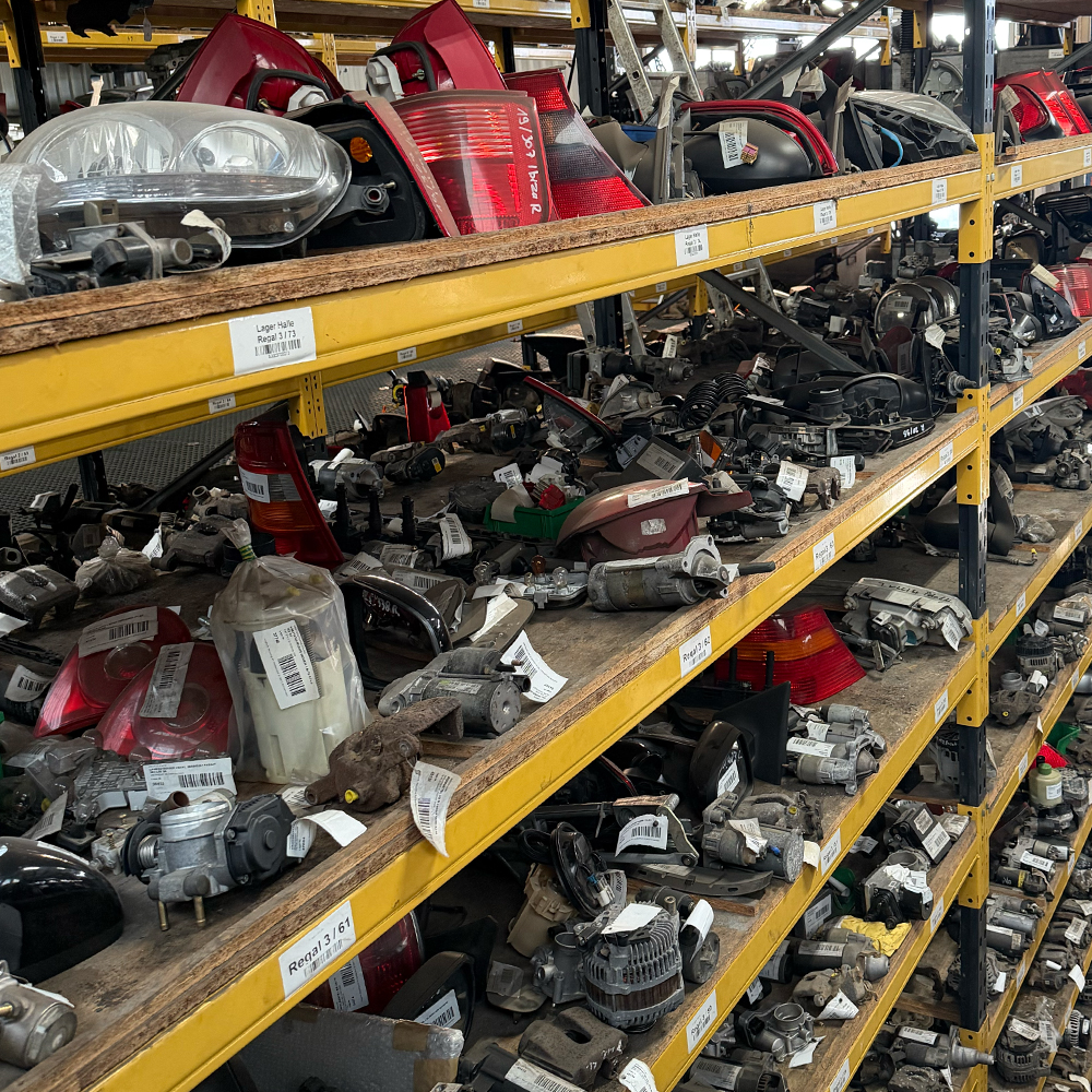 Ein gut sortiertes Lagerregal im Jungkurth eBay Shop, das dicht mit verschiedenen gebrauchten Auto-Ersatzteilen gefüllt ist. Auf den gelben Metallböden befinden sich unter anderem Scheinwerfer, Rückleuchten, Anlasser und Lichtmaschinen, die jeweils mit weißen Etiketten und Barcodes zur Identifizierung versehen sind.